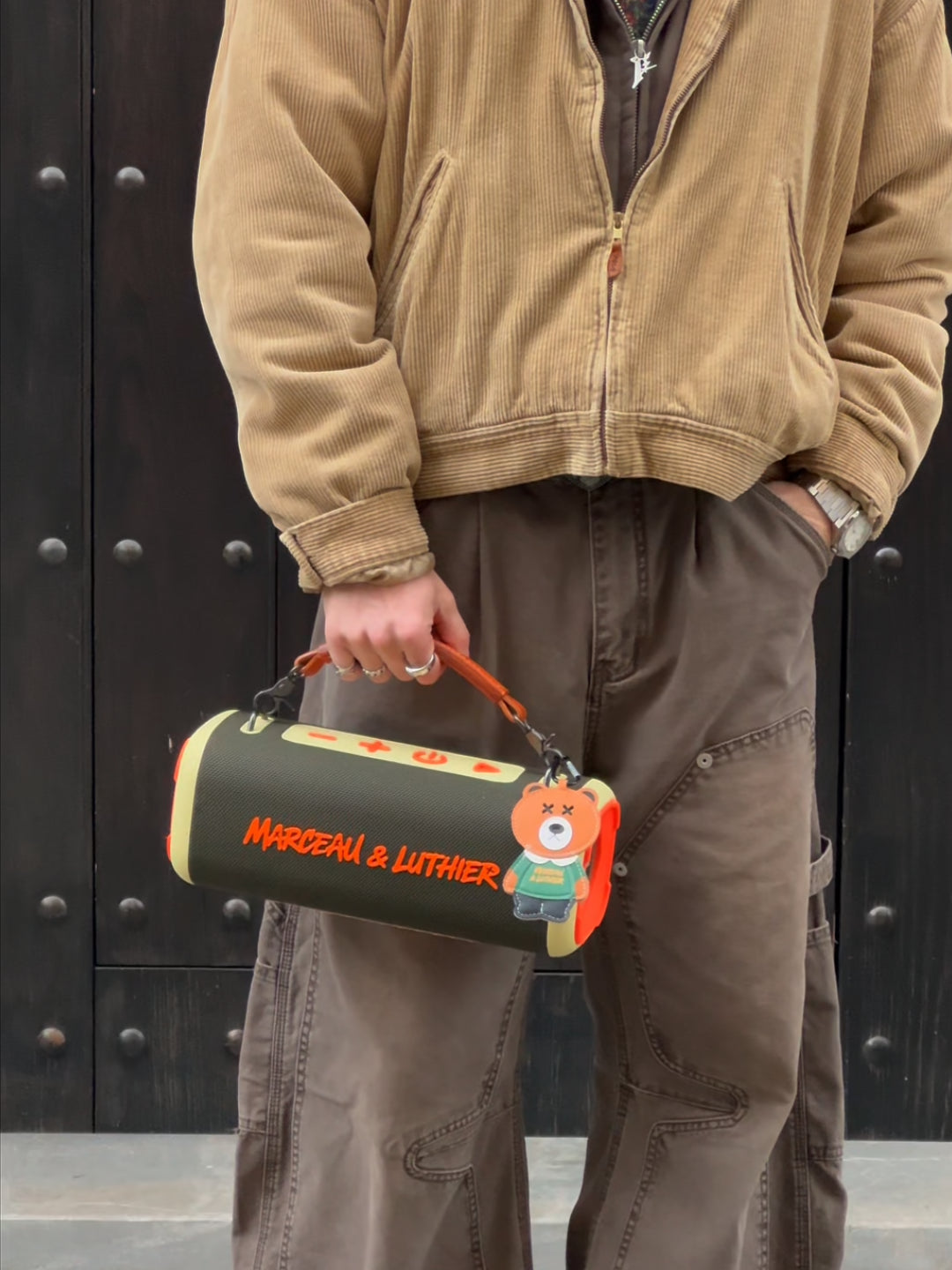 A close-up shot of a person holding the Nocturne speaker by its brown leather handle. The model is wearing a corduroy jacket and brown work pants. The speaker features orange "Marceau & Luthier" branding and a bear-shaped charm.