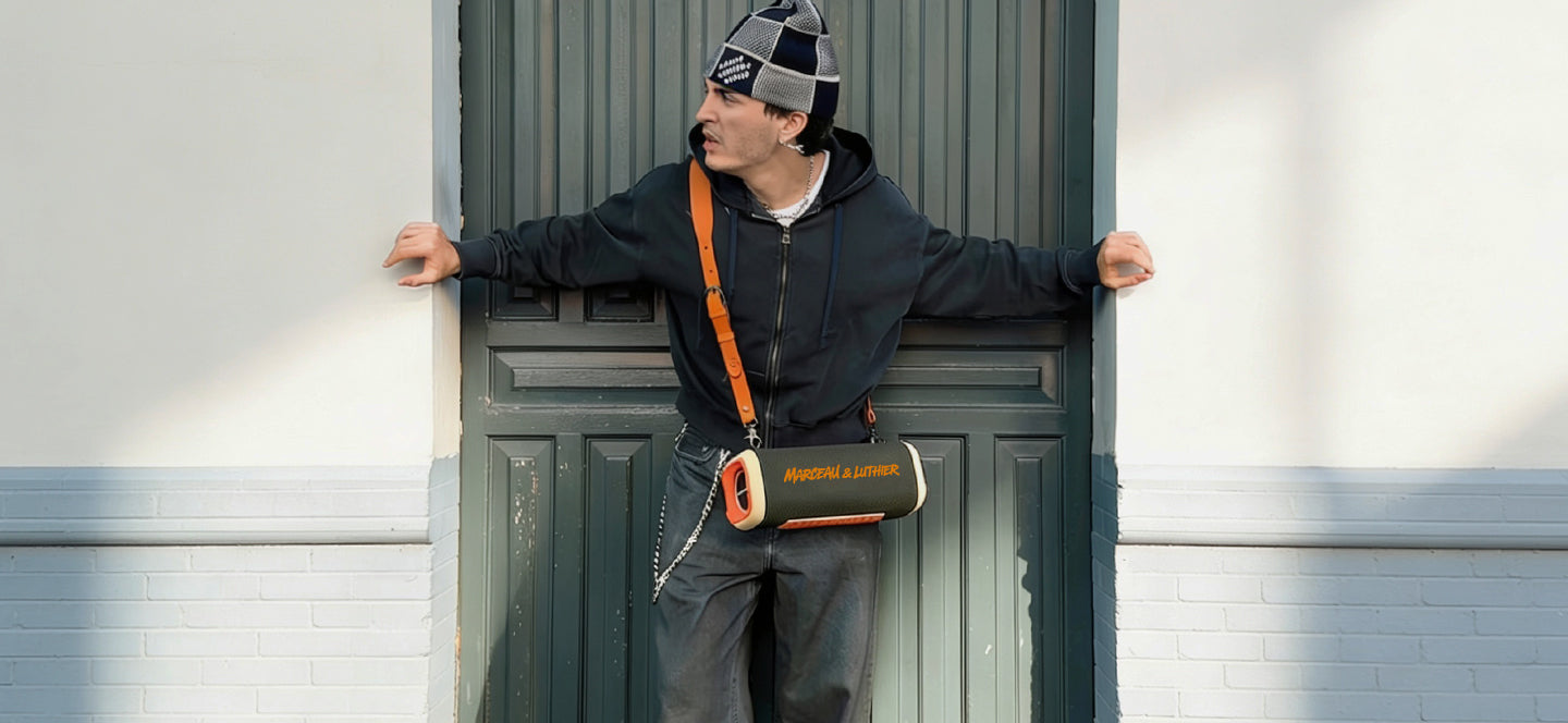 A man in streetwear clothing, a beanie, and a dark hoodie stands against a green wooden door, wearing the Marceau & Luthier NOCTURNE portable bluetooth speaker with its BANDOULIÈRE orange leather strap slung across his shoulder