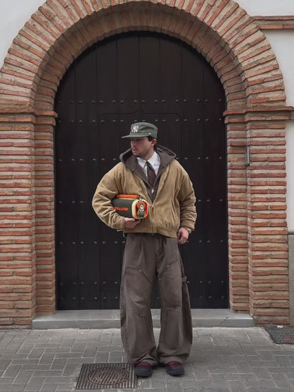 A video of a model standing in front of a large arched wooden door with brick detailing. He is wearing a tan corduroy jacket, a grey hoodie, a white shirt with a red tie, brown work pants, and a green cap. The model is holding the Nocturne portable speaker, which has an olive green body, orange branding, and a brown leather strap with a bear-shaped charm. The video shows him posing with the speaker under his arm and then walking away from the camera, carrying the speaker by its handle.