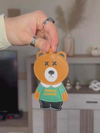 A video showcasing accessories for the Nocturne portable speaker. It begins with a person’s hand, wearing silver rings, holding up a brown leather bear charm with "Marceau & Luthier" printed on its green sweater. The video then shows an orange leather strap with the brand name embossed, followed by a close-up of a metal clasp and an embossed bear logo on the leather.