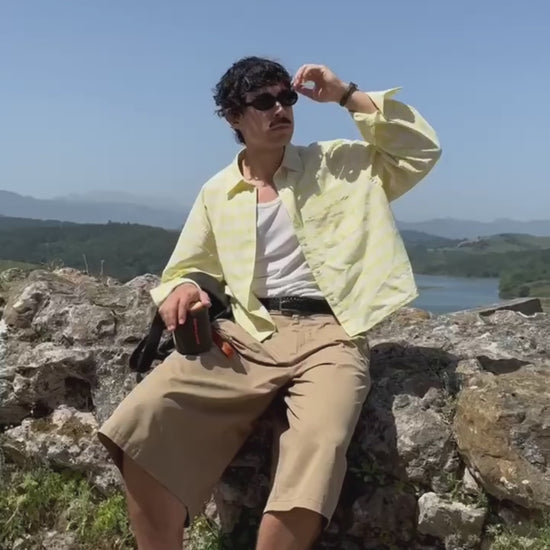 A man wearing a yellow plaid shirt and sunglasses sits on a rocky hill, holding a black and orange Marceau & Luthier BELLEVILLE portable bluetooth speaker. The video close-up shows the speaker's compact design and premium finish against a scenic landscape backdrop.
