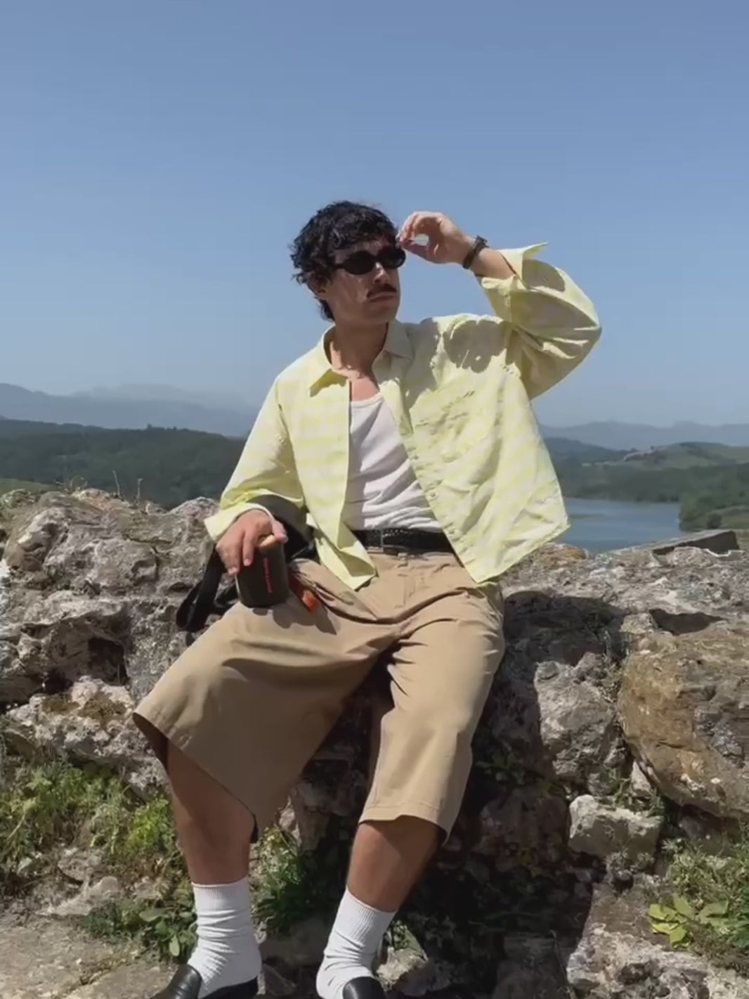 A man wearing a yellow plaid shirt and sunglasses sits on a rocky hill, holding a black and orange Marceau & Luthier BELLEVILLE portable bluetooth speaker. The video close-up shows the speaker's compact design and premium finish against a scenic landscape backdrop.